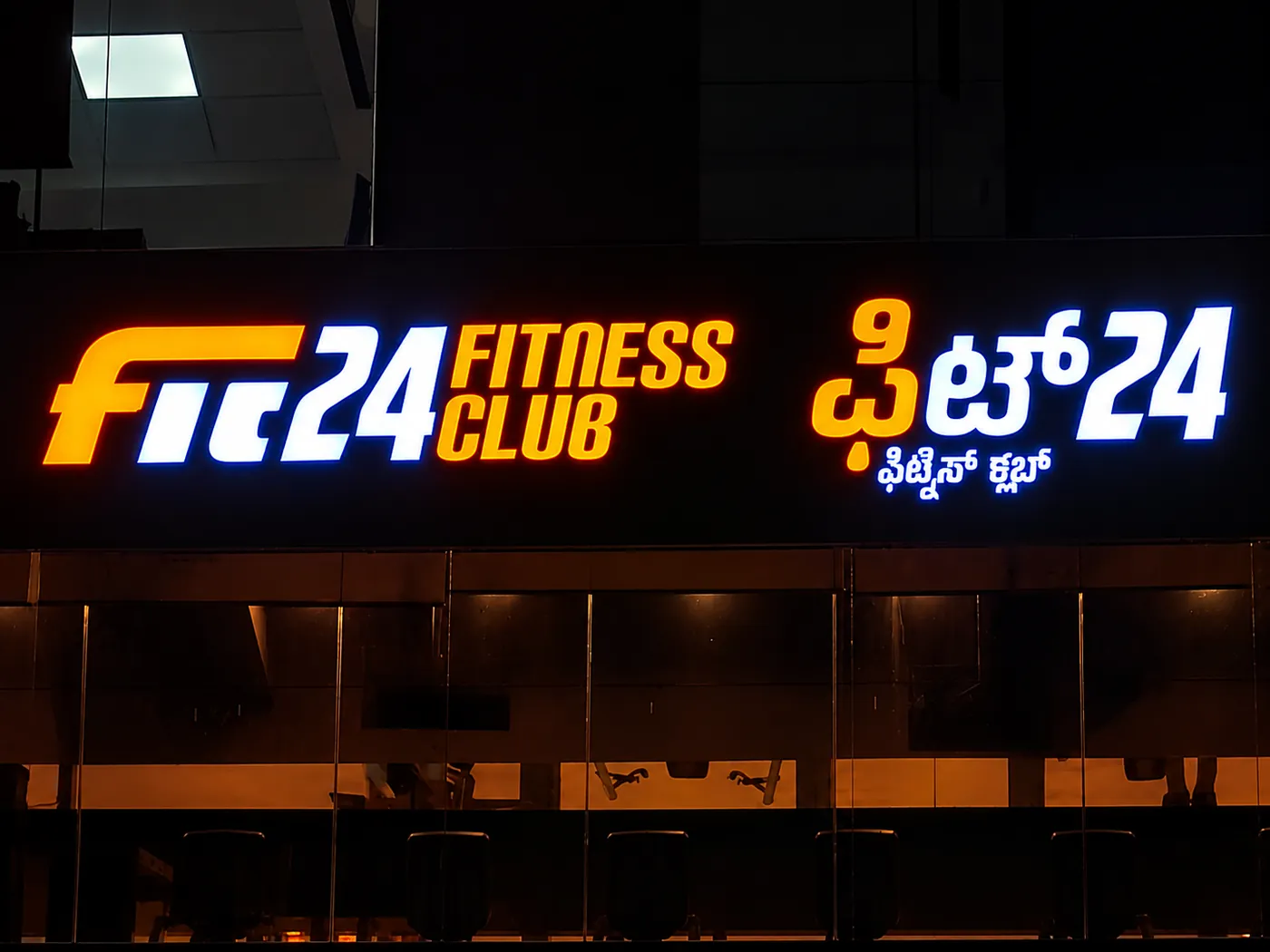 Fit24 Fitness Club backlit 3D sign board with orange and white letters and Kannada lettering on a dark facade at night